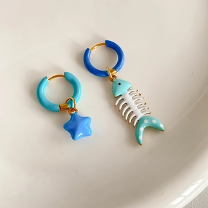 Detail of asymmetrical blue star and teal fishbone enamel earrings on white ceramic tray