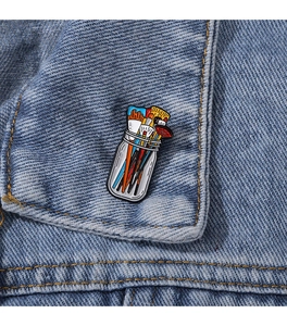 Detailed view of colorful paintbrush jar enamel pin pinned on a blue denim jacket pocket