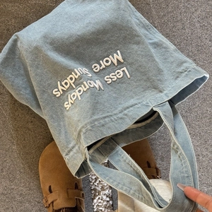 Close-up of light blue denim tote bag with white Less Mondays More Sundays embroidery
