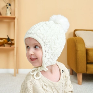 Close-up of white cable knit baby hat featuring a fluffy pompom and ear flaps with ties