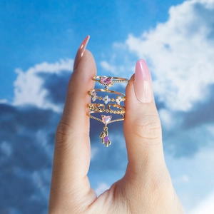 Detail of four gold rings with pink heart and bear charms held against a blue sky background