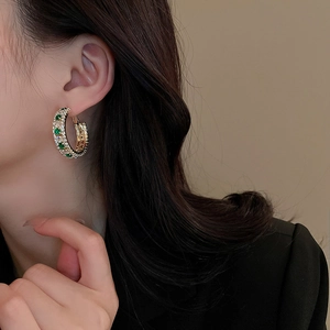 Golden C-shaped earrings with emerald and white cubic zirconia worn by a model
