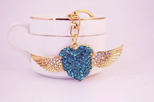 Detail of golden heart keychain with blue rhinestones and wing design against white cup