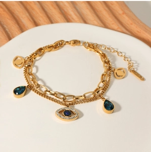 Close-up of gold layered bracelet with evil eye and blue teardrop stone on a white stand
