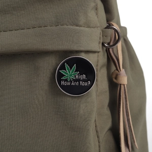 Detailed view of an enamel leaf pin with High How Are You text on a green canvas bag
