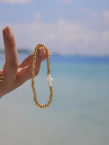 Detail of beaded gold bracelet with white cross charm held against a beach background