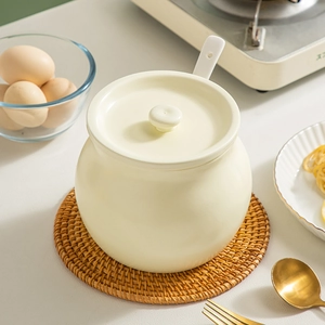 Creamy white ceramic lard pot with lid and spoon on a woven mat in a kitchen setting