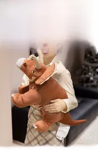 Close-up of a large brown triceratops plush pillow held by a person in a cozy room