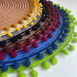 Round woven placemats with pom-pom fringe in multiple metallic colors stacked on white