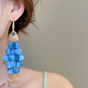 Close-up of golden fish-tail earrings with blue scales worn by a model in soft light