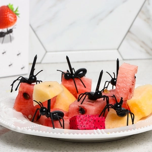 Black ant-shaped fruit picks used on fresh fruit pieces in a white round plate