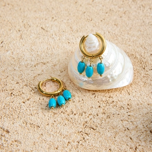 Detail of 18k gold earrings featuring three turquoise drops on a seashell and sand background