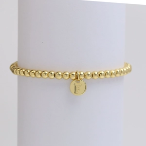 Detail of a gold beaded bracelet featuring a round charm with the letter F engraving