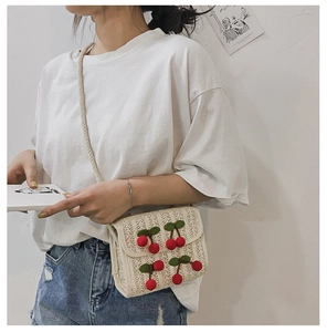 Detail of beige straw crossbody bag featuring red cherry pom-poms and woven strap