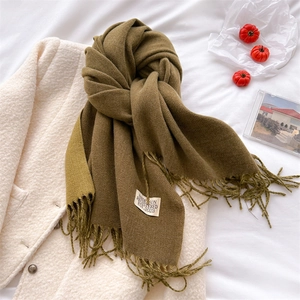 Detail view of olive and tan cashmere-feel scarf with fringe and brand label on white bed