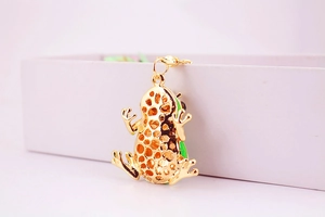Detail of a golden frog-shaped metal earring with green enamel finish on a white box