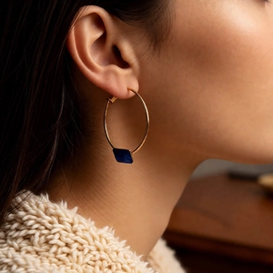 Macro detail of gold-plated hoop earrings featuring a square blue gemstone on a model