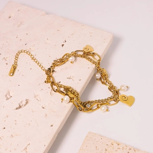 Layered gold titanium bracelet with white pearls and heart pendants on stone surface