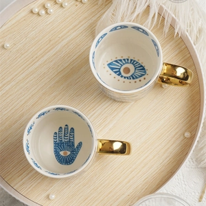 Ceramic evil eye coffee mugs with gold handles on a rustic tray with pearls and feathers