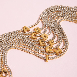 Detail van gouden tennisarmbanden met strass letters A-I op een zachte roze achtergrond