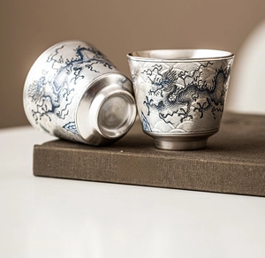 Detalle macro de tazas de té plateadas con patrón de dragón sobre un libro antiguo