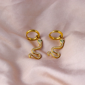 Macro shot of gold snake earrings with white zircons and green eyes on pink silk fabric