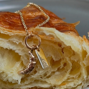 Macro detail of gold croissant pendant and OUI tag on necklace atop fresh bread