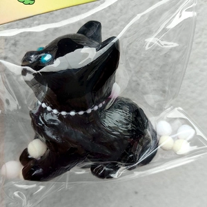 Close-up of a squishy black cat stress toy with blue eyes and pearl necklace in packaging