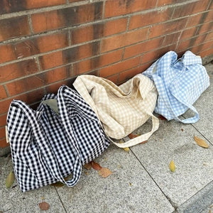 Three large plaid canvas tote bags in black, beige, and blue leaning against a brick wall