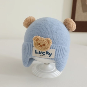 Close-up of knit baby hat with double pom poms and a lucky bear patch in sky blue