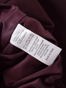 Detailaufnahme des Pflegeetiketts und Goldknopfes an einer burgunderroten Polyesterjacke