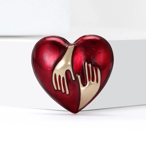 Deep red enamel heart brooch featuring gold-plated hugging hands on white geometric stand