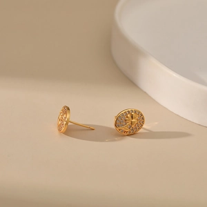 Oval gold earrings with clover engraving and zircon accents on a warm beige background