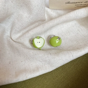 Detailed view of green apple stud earrings with white enamel slice on beige linen