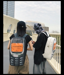 Two people wearing retro mobile phone shaped backpacks in grey and white on a city bridge