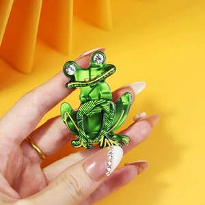 Handheld view of a green acrylic frog brooch with gold outlines and sparkling rhinestone eyes