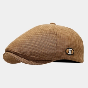 Detail of brown plaid newsboy cap featuring a round leather patch with an m letter tag