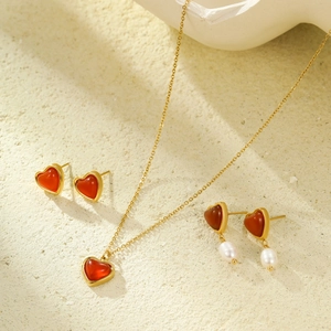 Golden heart necklace and pearl earrings jewelry set with red stones on sand background