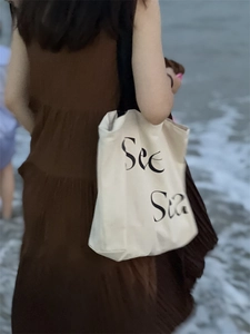 Minimalist white canvas tote bag with See Sea print shown against ocean waves