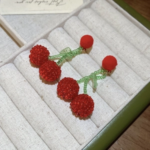 Detailed view of handmade beaded cherry earrings with red pom-poms on white velvet display