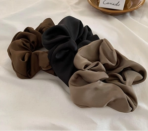 Close-up of three satin scrunchies in black, brown, and khaki on a soft fabric background