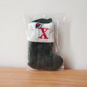 Green knit Christmas stocking in packaging featuring red letter X and Santa hat embroidery