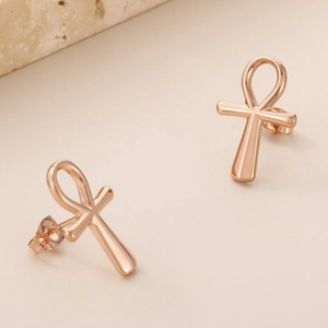 Macro shot of 18K rose gold plated Ankh cross stud earrings on a warm beige background