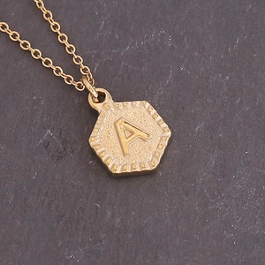 Minimalist gold hexagon necklace with embossed initial A pendant on a textured dark background