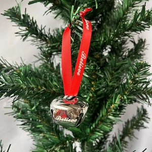 Close-up of a silver Christmas bell with red ribbon printed with Believe on a pine tree