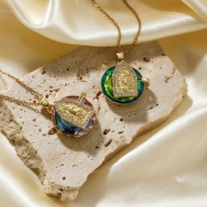 Gold Virgin Mary pendant necklace with colorful crystal base on beige natural stone background