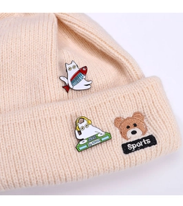 Nahaufnahme einer beigen Strickmütze mit Katzen-Emaille-Pins und kleiner Bären-Stickerei