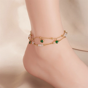 Detail of a layered gold anklet featuring green heart charms and white crystal beads