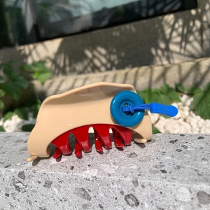 Minimalist beige hair claw clip with red teeth and blue button detail on a stone surface