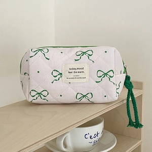 Quilted white cotton makeup bag with green bow patterns and zipper on a wooden shelf